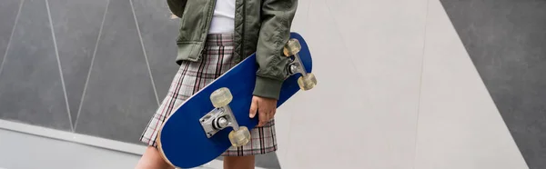 Vista recortada de la chica preadolescente con estilo en chaqueta de bombardero y falda que sostiene penny board cerca del centro comercial, pancarta - foto de stock