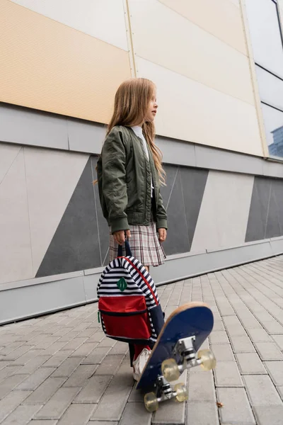 Full length of stylish preteen girl in bomber jacket holding backpack while riding penny board near mall — Stockfoto