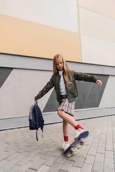Full length of preteen girl in stylish bomber jacket holding backpack while riding penny board near mall - foto de stock