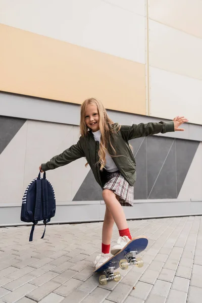 Full length of happy preteen girl in stylish bomber jacket holding backpack while riding penny board near mall - foto de stock
