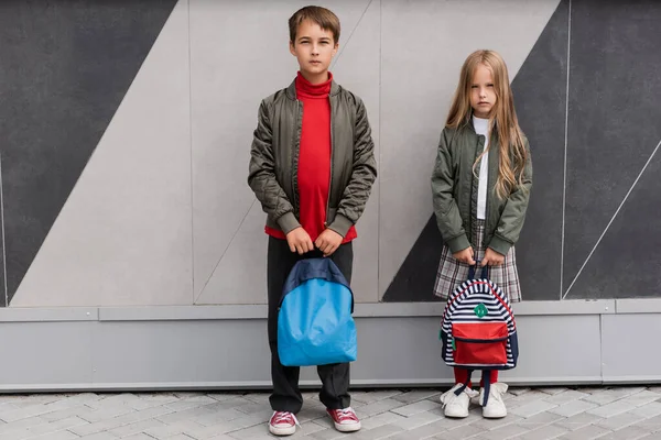 Полная длина стильные дети в куртках бомбардировщика держа рюкзаки, стоя рядом с торговым центром — стоковое фото