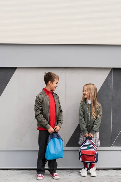 Full length of happy and well dressed kids in bomber jackets holding backpacks while standing near mall - foto de stock