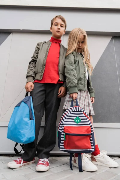 Low angle view of stylish kids in bomber jackets holding backpacks while standing near mall — Fotografia de Stock
