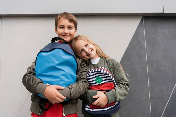 Низький кут зору щасливих дітей в модних куртках бомбардувальника, що тримають рюкзаки, стоячи біля торгового центру — стокове фото