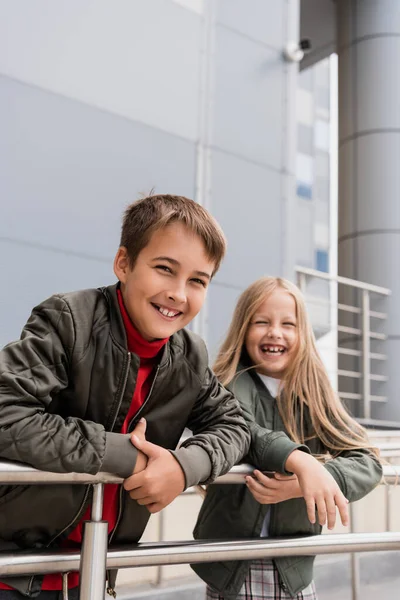 Cheerful preteen kids in bomber jackets leaning on metallic handrails near mall — Stockfoto