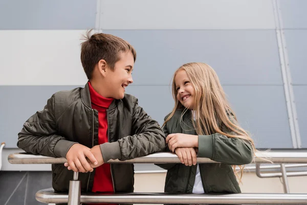Happy preteen kids in bomber jackets looking at each other while leaning on metallic handrails near mall — Stock Photo