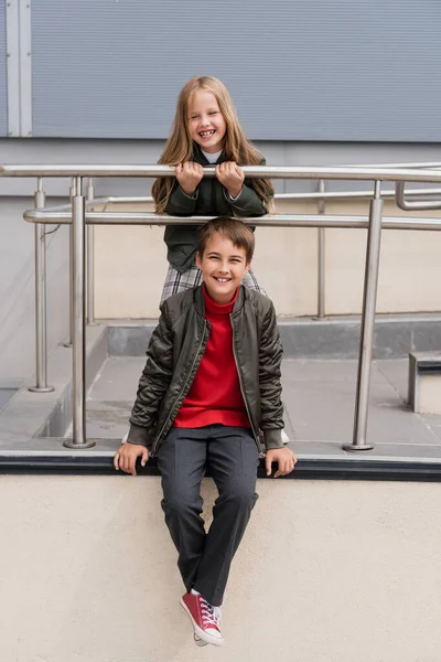 Happy preteen kids in stylish bomber jackets posing near metallic handrails near mall — Photo de stock