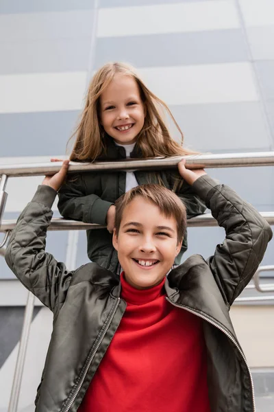 Blick auf glückliche und gut gekleidete Kinder in Bomberjacken, die sich an Metallgeländer in der Nähe des Einkaufszentrums lehnen — Stockfoto