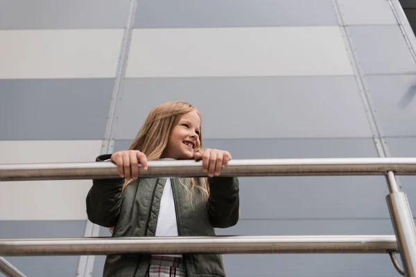 Low angle view of cheerful preteen girl in bomber jacket leaning on metallic handrails near mall - foto de stock