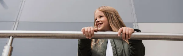 Low angle view of cheerful and well dressed preteen girl in bomber jacket leaning on metallic handrails near mall, banner — Fotografia de Stock