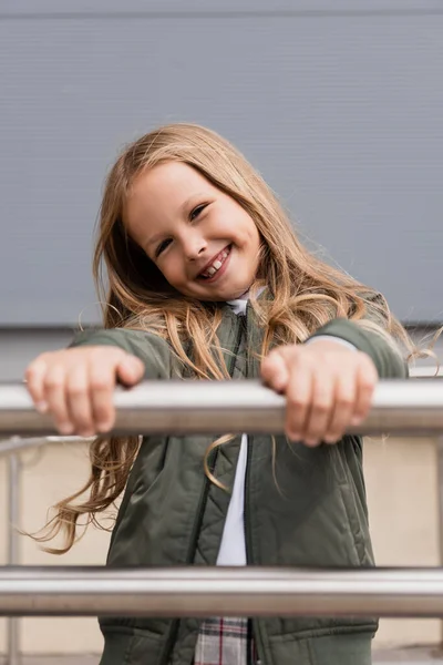 Cheerful preteen girl in stylish bomber jacket leaning on metallic handrails near mall — Stock Photo