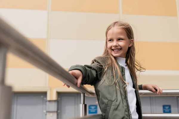 Satisfied preteen girl in stylish bomber jacket leaning on metallic handrails near mall — Fotografia de Stock