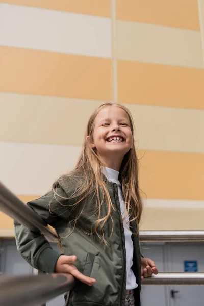 Visão de baixo ângulo da menina satisfeita na jaqueta bomber elegante inclinando-se em corrimãos metálicas perto do shopping — Fotografia de Stock