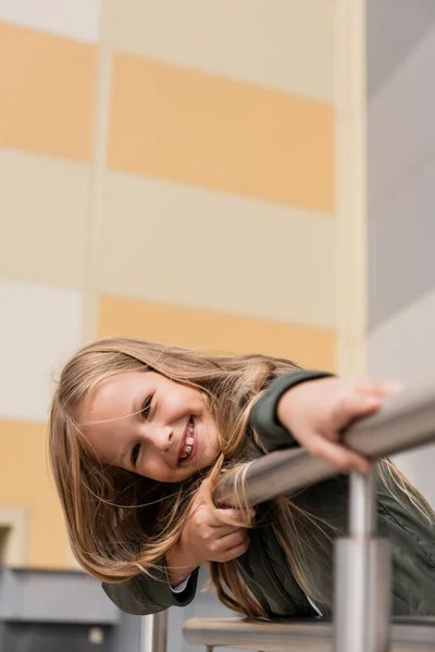 Carefree girl in stylish bomber jacket leaning on metallic handrails near mall — Photo de stock