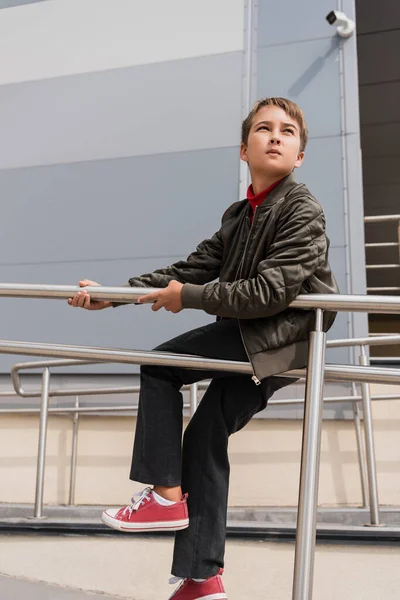 Preteen boy in stylish bomber jacket sitting on metallic handrails near mall — Photo de stock