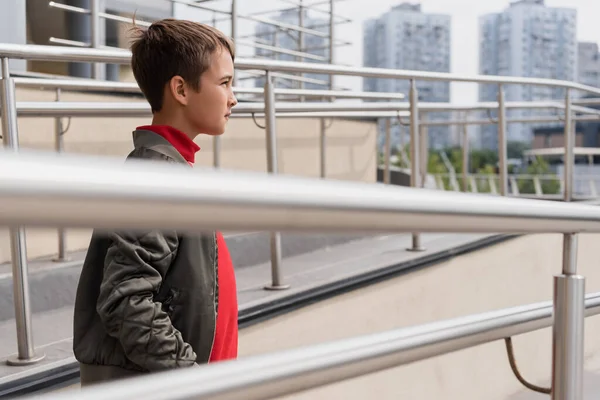 Well dressed preteen boy in stylish bomber jacket standing near metallic handrails on blurred foreground — Photo de stock