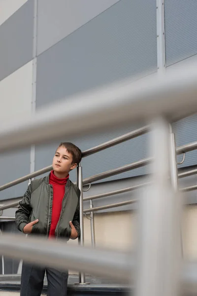Well dressed preteen boy in stylish bomber jacket posing with hands in pockets near metallic handrails on blurred foreground — Photo de stock