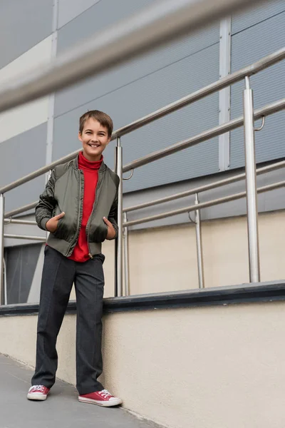 Happy preteen boy in stylish bomber jacket posing with hands in pockets near metallic handrails on blurred foreground — Foto stock