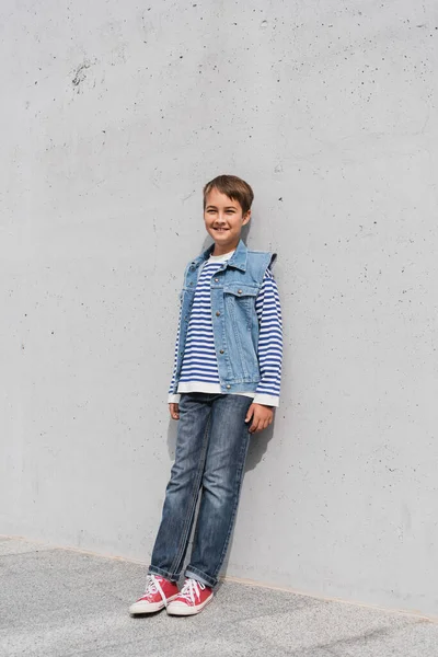 Pleine longueur de garçon heureux en chemise à manches longues rayée et gilet en denim appuyé sur le mur à l'extérieur — Photo de stock