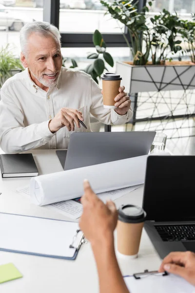 Positive middle aged businessman holding coffee to go near laptops and african american colleague in office — Stock Photo