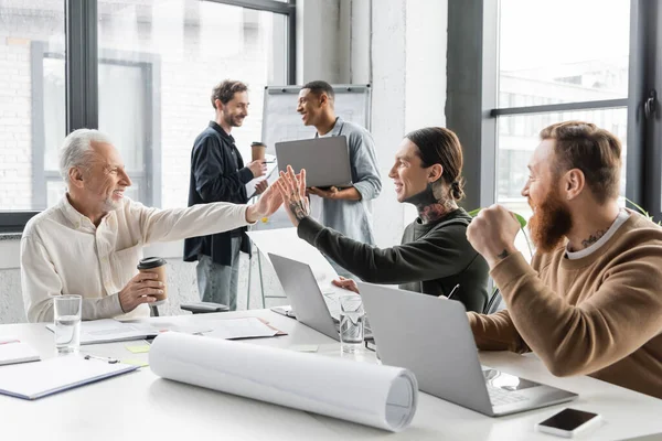 Empresários alegres dando alta cinco perto de dispositivos e papéis durante a reunião no escritório — Fotografia de Stock