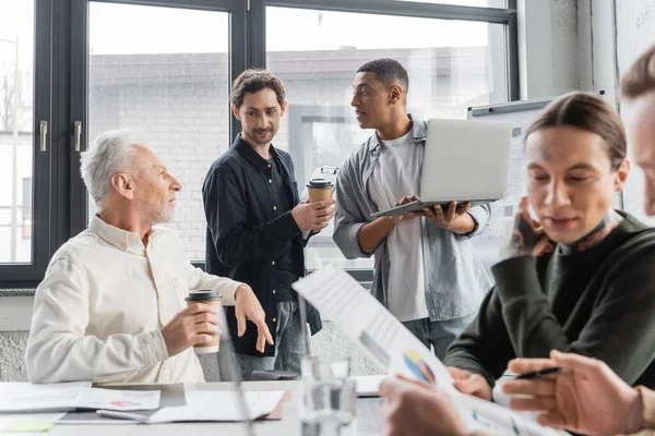 Мультиэтнические бизнесмены с кофе и ноутбуком разговаривают во время встречи в офисе — стоковое фото
