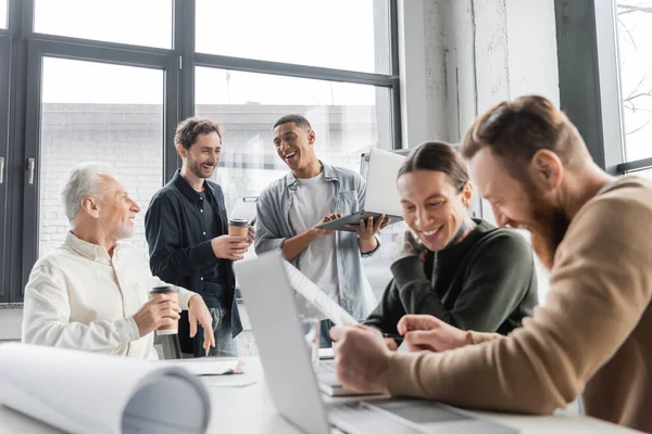Empresários inter-raciais alegres com café para ir e laptop conversando durante reunião no escritório — Fotografia de Stock