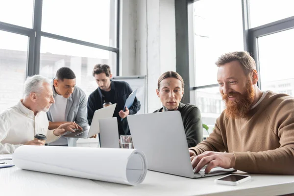 Uomo d'affari sorridente che utilizza laptop vicino ai colleghi durante la riunione di lavoro in ufficio — Foto stock