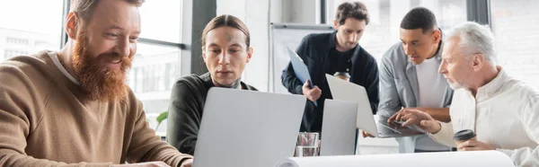 Позитивний бізнесмен використовує ноутбук біля татуйованого колеги в офісі, банер — стокове фото