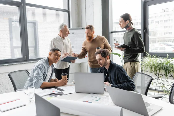 Мультиэтнические деловые люди с кофе идти с помощью ноутбука рядом с бумажной работой в офисе — стоковое фото