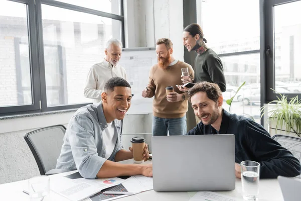 Позитивні багатоетнічні бізнесмени використовують ноутбук біля колег під час зустрічі в офісі — стокове фото