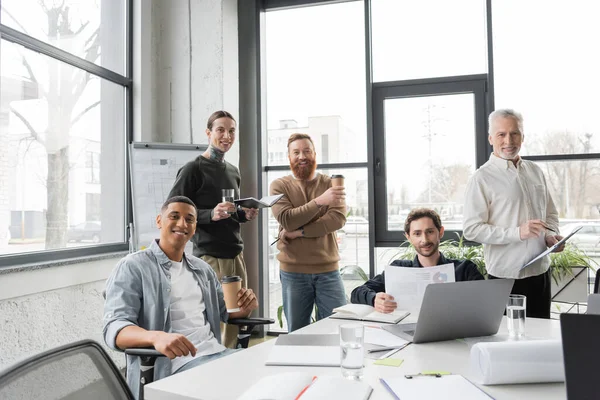 Des hommes d'affaires multiethniques positifs avec des papiers et des ordinateurs portables regardant la caméra lors d'une réunion au bureau — Photo de stock