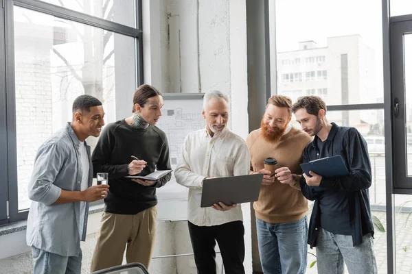 Smiling mature businessman using laptop near interracial colleagues during presentation in office — Stock Photo