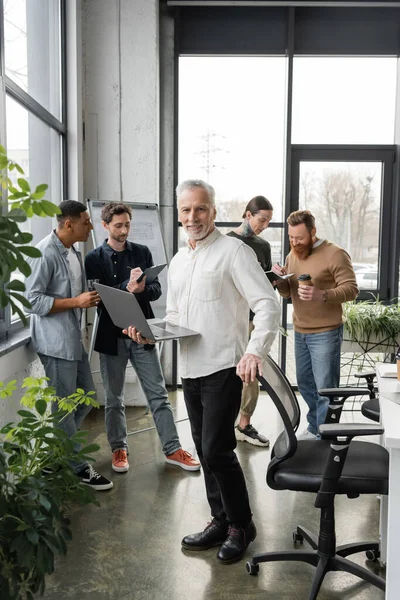 Positiver Geschäftsmann mittleren Alters hält Laptop während eines Treffens im Büro neben interrassischen Kollegen — Stockfoto