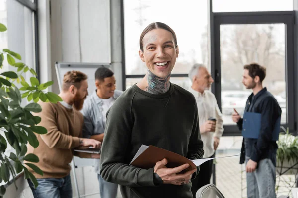 Cheerful tattooed businessman holding paper folder and looking at camera near interracial colleagues in office — Stock Photo