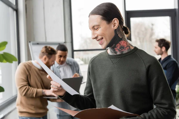 Homme d'affaires tatoué souriant tenant un dossier papier près de collègues multiethniques flous au bureau — Photo de stock
