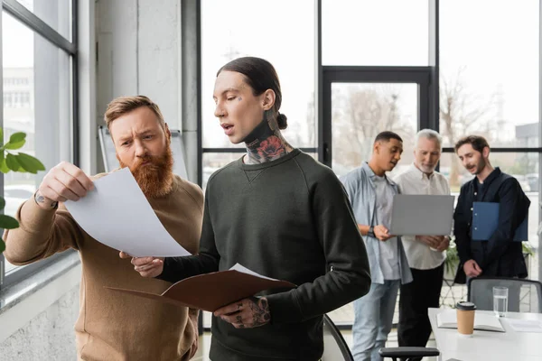 Des hommes d'affaires tatoués travaillant avec des journaux proches de collègues interraciaux flous au bureau — Photo de stock