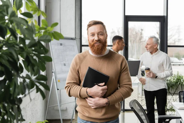 Empresário sorridente segurando caderno perto de plantas borradas e colegas multiétnicos no escritório — Fotografia de Stock