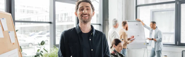 Усміхнений бізнесмен дивиться на камеру біля дошки і розмиті колеги в офісі, банер — стокове фото