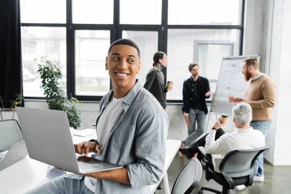 Joyeux homme d'affaires afro-américain utilisant un ordinateur portable près de collègues flous travaillant dans le bureau — Photo de stock
