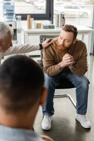 Middle aged man calming tattooed person with alcohol addiction during therapy in rehab center — Stock Photo