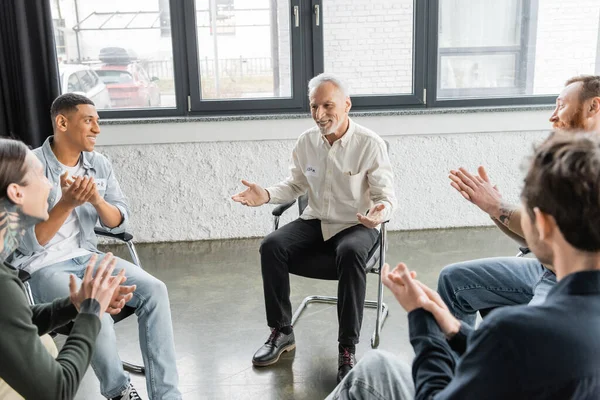Positive mature man with alcohol addiction sitting in circle while multiethnic people applauding in rehab center — Stock Photo