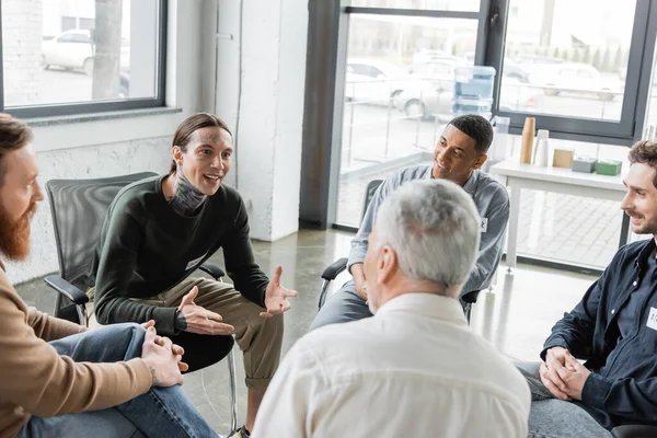 Cheerful tattooed man with alcohol addiction talking to interracial group during therapy in rehab center — Stock Photo