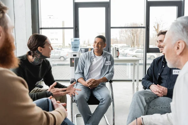 Tattooed man with alcohol addiction talking to multiethnic group in rehab center — Stock Photo