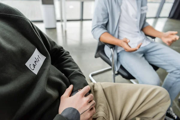 Cropped view of man with name sticker sitting in circle during alcoholics meeting in rehab center — Stock Photo