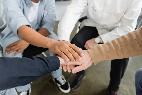 Vista cortada de pessoas inter-raciais sentadas em círculo e colocando as mãos juntas durante a reunião de alcoólicos no centro de reabilitação — Fotografia de Stock