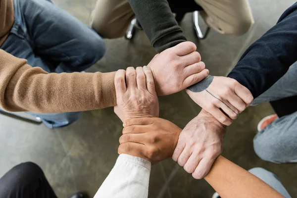 Top view of multiethnic people with alcohol addiction joining hands during group therapy in rehab center — Stock Photo