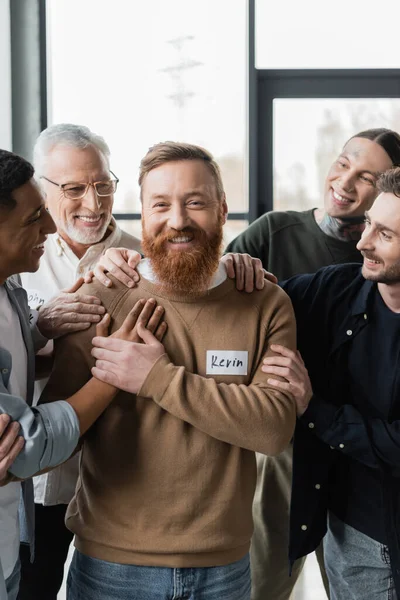 Personas multiétnicas positivas que abrazan al hombre durante la terapia grupal con alcohólicos en el centro de rehabilitación - foto de stock