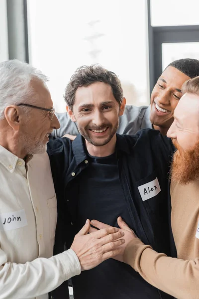Interracial people hugging cheerful man during alcoholics therapy in rehab center — Stock Photo