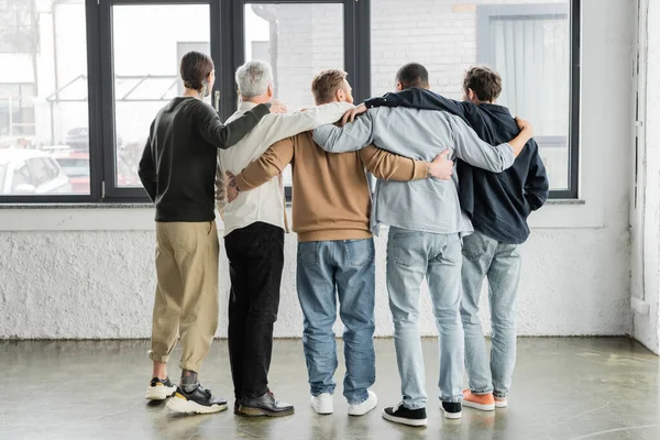 Vista trasera de hombres interracial abrazándose durante reunión anónima de alcohólicos en el centro de rehabilitación - foto de stock